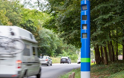 Un stâlp cilindric albastru înalt, prevăzut cu camere de supraveghere și senzori, amplasat pe marginea unei șosele mărginite de copaci în timp ce trec vehicule.