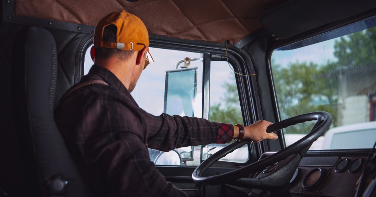 Imaginea prezintă un șofer purtând șapcă aflat la volanul unui camion, privind spre oglinda laterală în timp ce conduce.