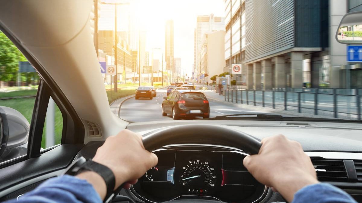 Șofer aflat la volanul unei mașini aflate în trafic. Șoferul ține ambele mâini pe volan. În față se observă mai multe vehicule pe o șosea cu două benzi, clădiri moderne de birouri și indicatoare rutiere