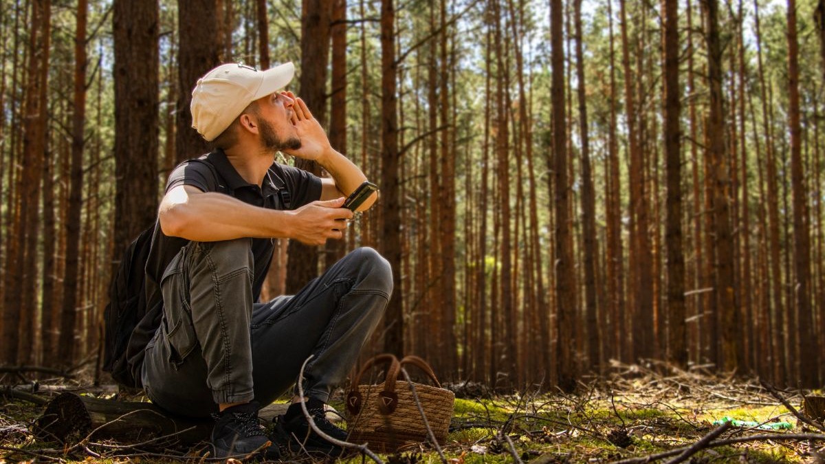 Tânăr îmbrăcat cu blugi gri și tricou negru, cu șapcă crem pe cap, cu un telefon în mână, în timp ce stă pe un lemn, într-o pădure și strigă după ajutor