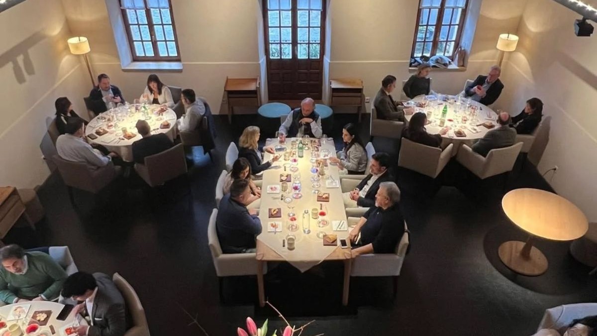 Această fotografie surprinde de sus interiorul unui restaurant spațios, cu pereți de un alb mat și o podea închisă la culoare, unde mai multe grupuri de oameni sunt așezate la mese. În centrul compoziției se află o masă dreptunghiulară lungă, flancată de alte patru mese rotunde dispuse simetric spre colțurile încăperii, toate fiind acoperite cu fețe de masă albe și setate cu pahare, farfurii mici și tacâmuri.