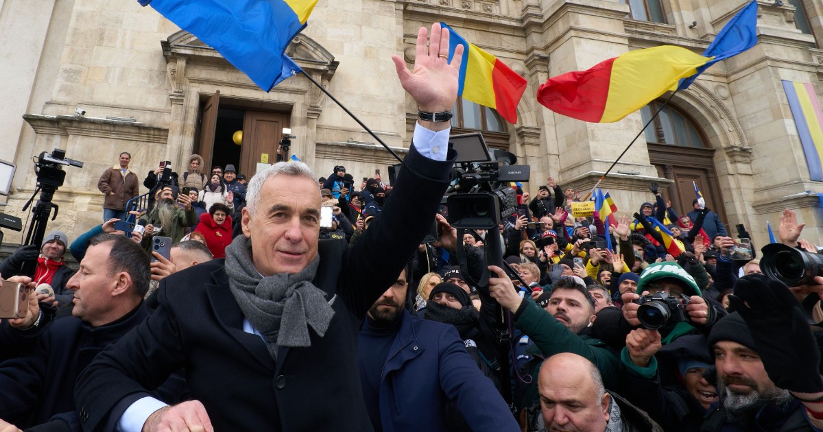 În imagine se vede un bărbat care salută mulțimea, înconjurat de susținători și jurnaliști, în fața unei clădiri oficiale. Oamenii din jur flutură steaguri tricolore și filmează momentul.