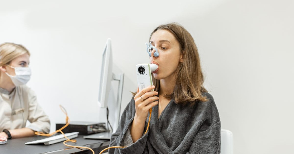 O femeie sta intr-un cabine tpe scaun si sufla in spirometru, iar un medic in fata ei urmareste testul. Varsta plamanilor