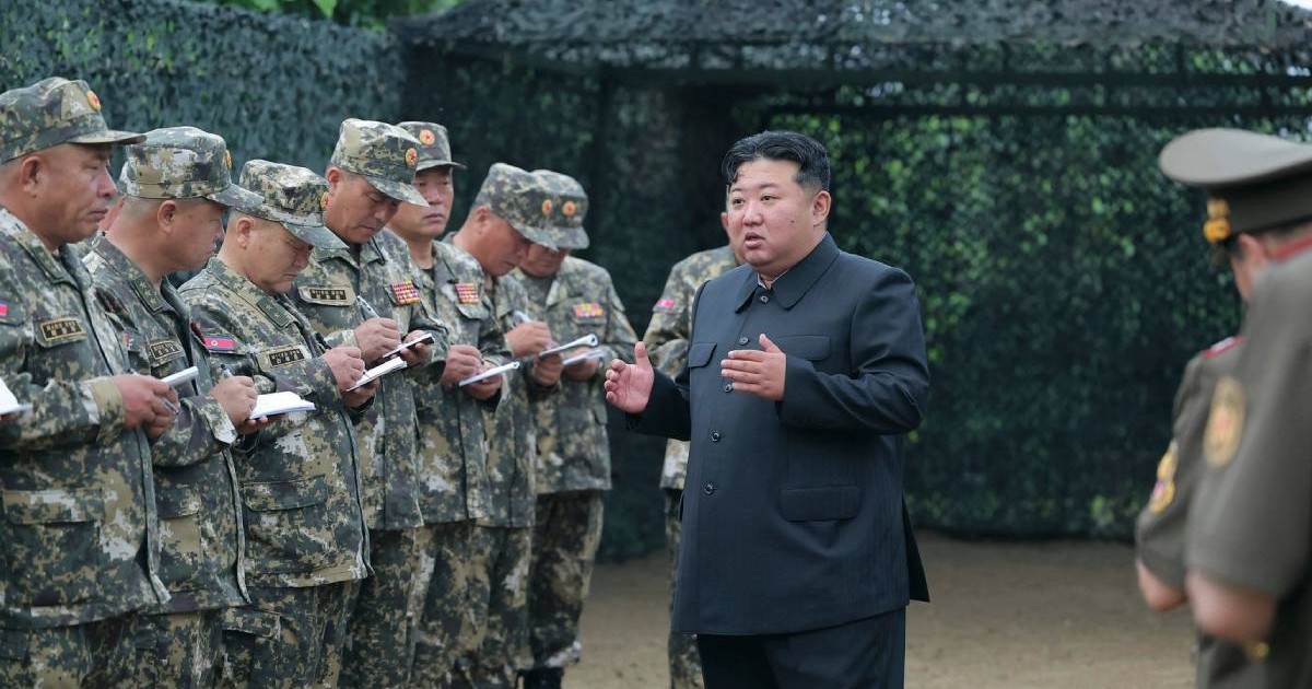Kim Jong Un alături de soldații săi, în timp ce le vorbește. Poartă un costum negru iar militarii au uniforme