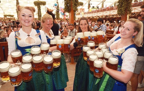 Mai multe chelnerițe îmbrăcate în costume tradiționale bavareze purtând numeroase halbe mari pline cu bere în interiorul unui cort aglomerat de la Oktoberfest.