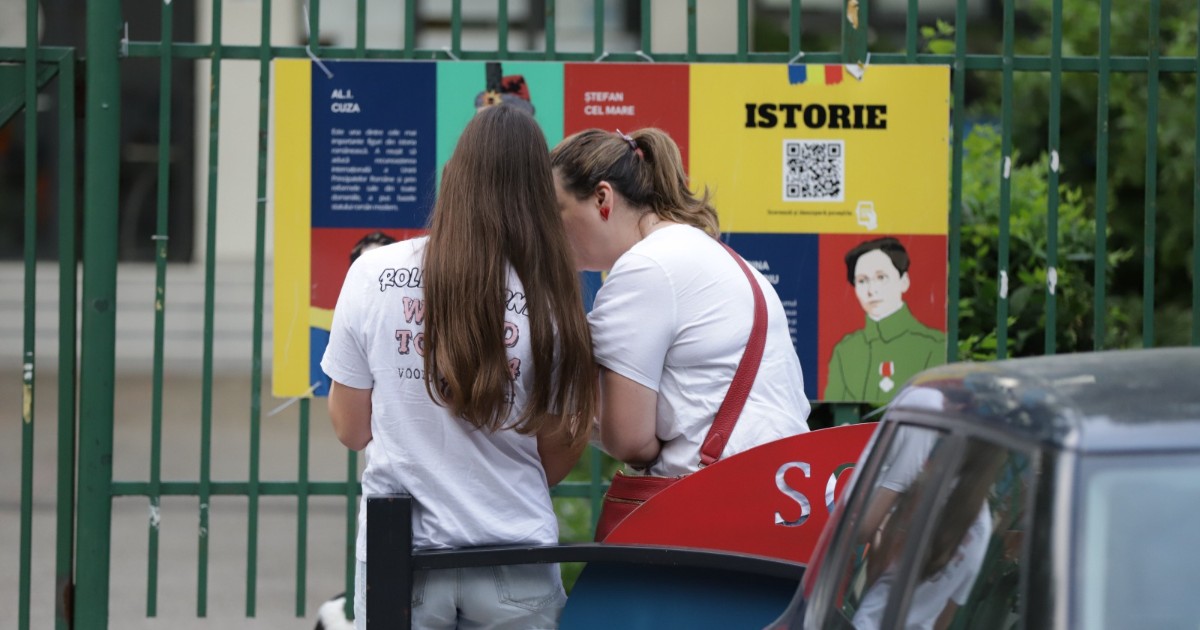 Două tinere, privite din spate, îmbrăcate în tricouri albe, care stau de vorbă în fața unui panou educațional colorat, montat pe un gard metalic verde.