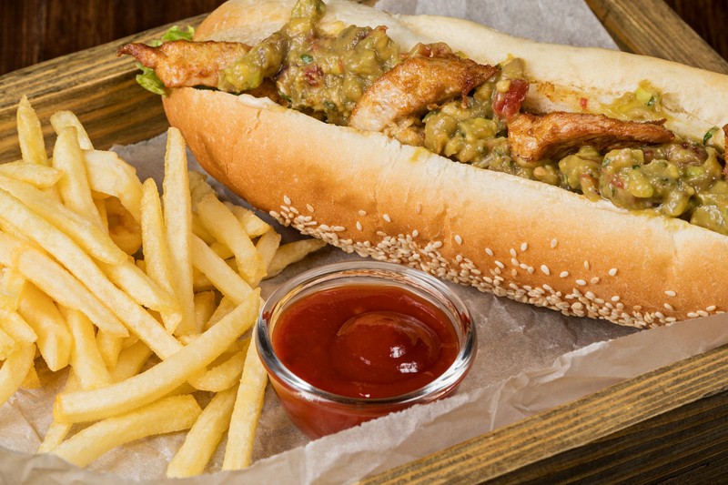 Hot dog with grilled chicken breast, french fries and sauce on paper in a wooden box