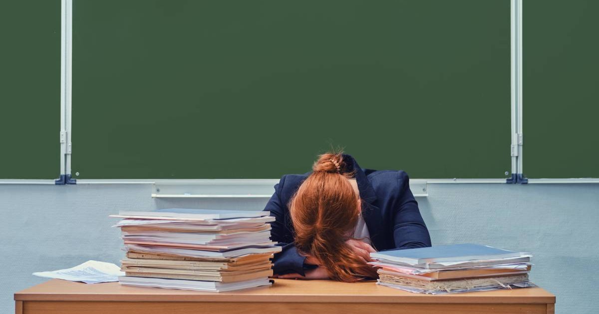 O profesoară care stă cu capul pe catedră, are fața acoperită și părul roșcat, este supărată și înconjurată de cărți