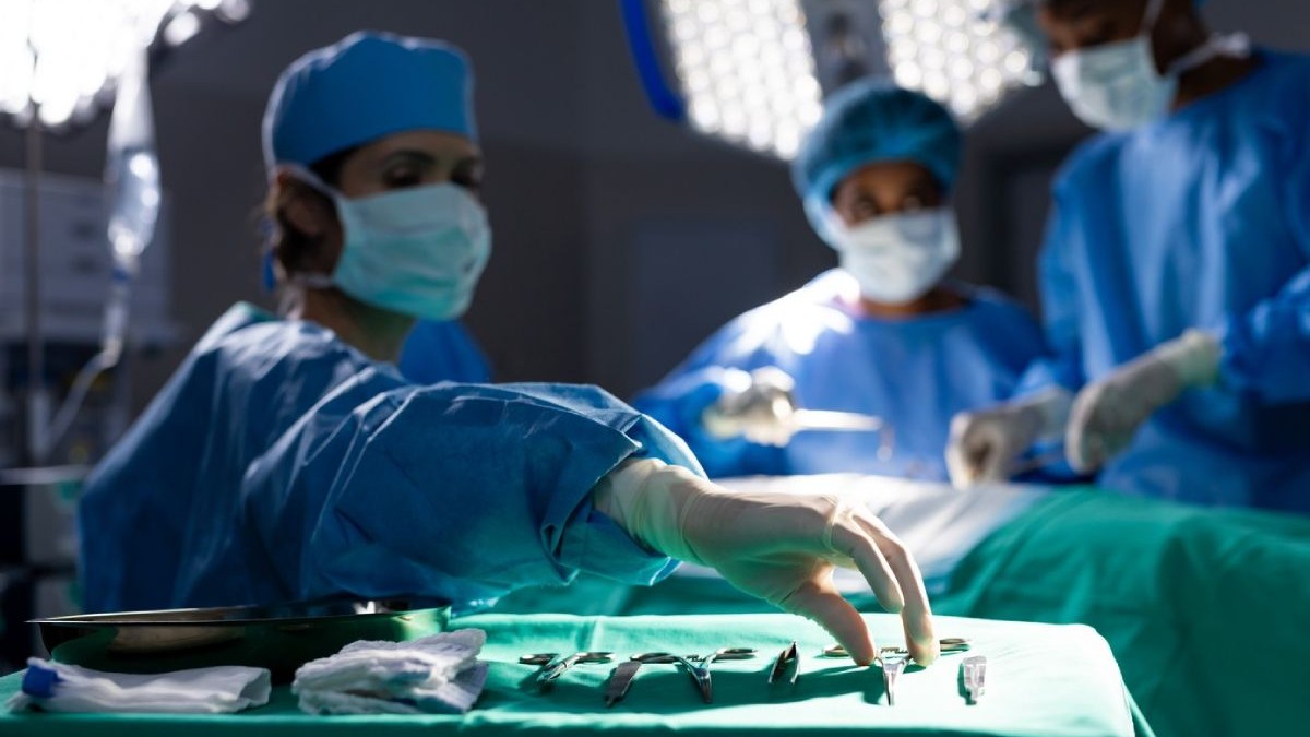 Trei medici în costume sterile albastre, cu masti pe față in timpul unei operații, in afat se vede instrumentarul steril