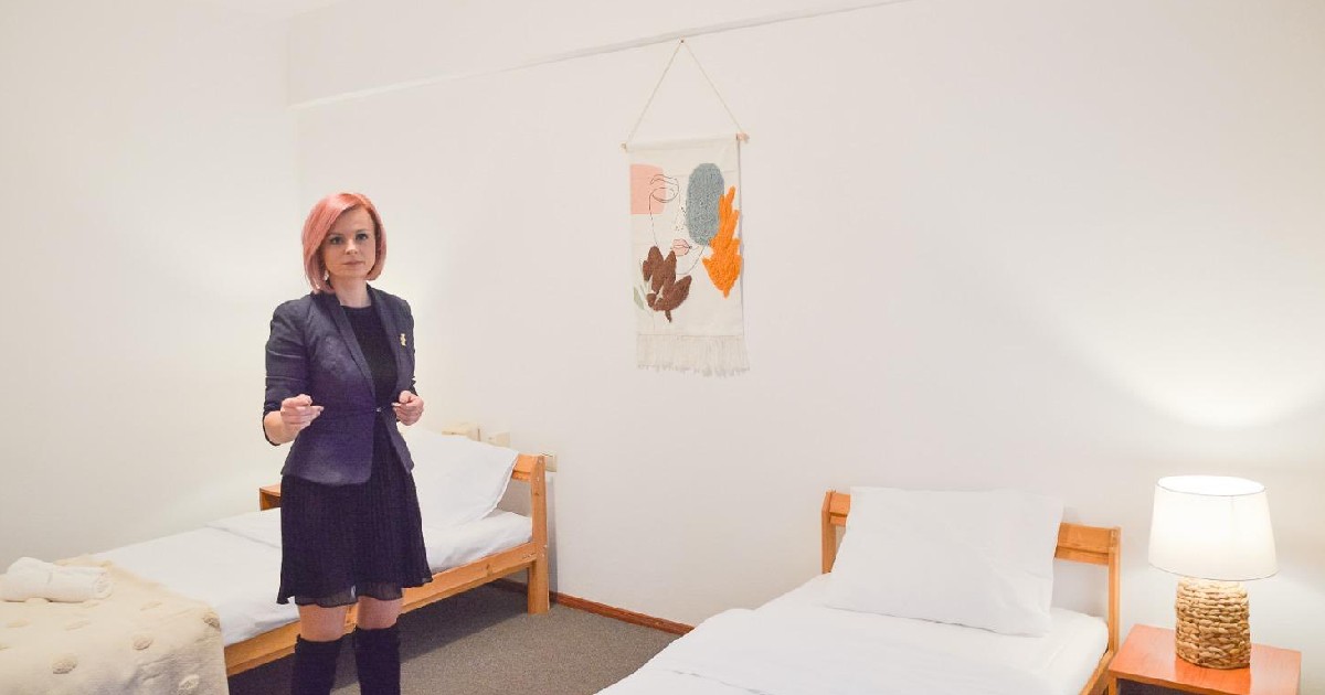 Flavia Boghiu îmbrăcată într-o rochie neagra și un sacrou gri închis, aflându-se într-o cameră din adăpost