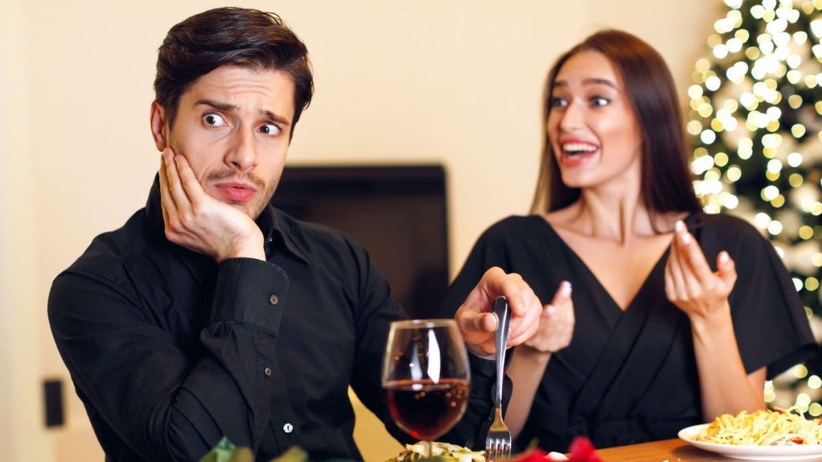 Un cuplu tânăr la o întâlnire într-un restaurant; bărbatul are o expresie amuzantă de dezamăgire și șoc, se simte stânjenit în timp ce ascultă o femeie entuziasmată vorbind.