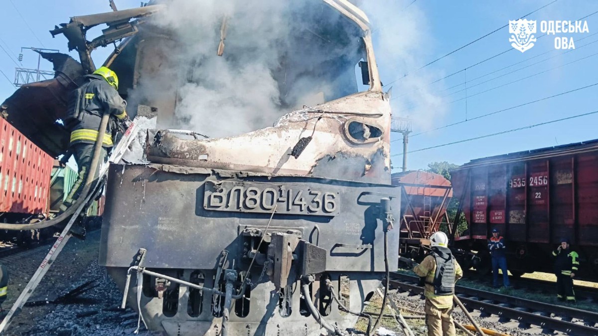 Locomotiva unui tren apare grav avariată și cuprinsă de fum, după atac, în timp ce pompierii intervin pentru stingerea incendiului. În jurul locomotivei se văd resturi și echipamente deteriorate, iar mai multe vagoane sunt oprite pe liniile de cale ferată.