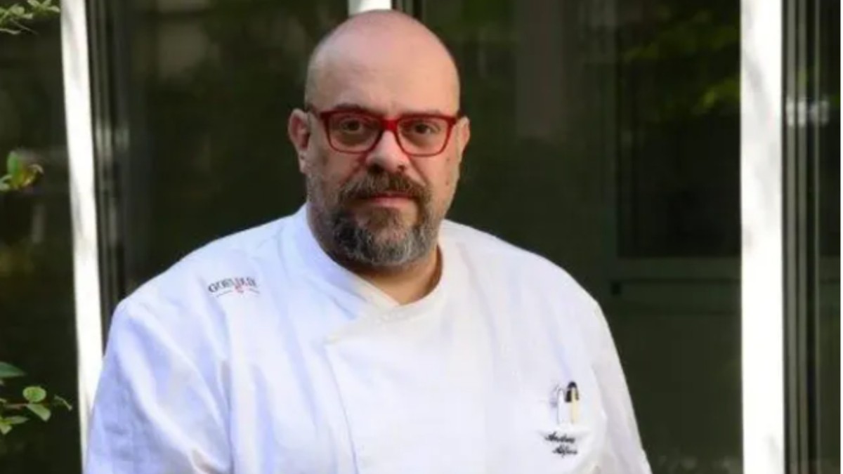 Imaginea îl prezintă pe un bucătar îmbrăcat în uniformă albă profesională, cu ochelari cu rame roșii, stând în fața unei clădiri cu uși din sticlă. Aspectul său sobru și postura calmă sugerează experiență și autoritate într-un context profesional din domeniul gastronomiei.