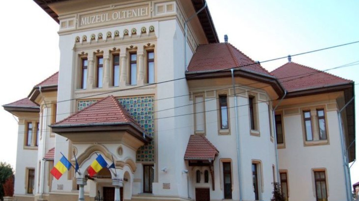Imaginea prezintă clădirea Muzeului Olteniei din Craiova. Fațada muzeului este bej, iar acoperișul este din țiglă roșie. La intrare se observă patru steaguri