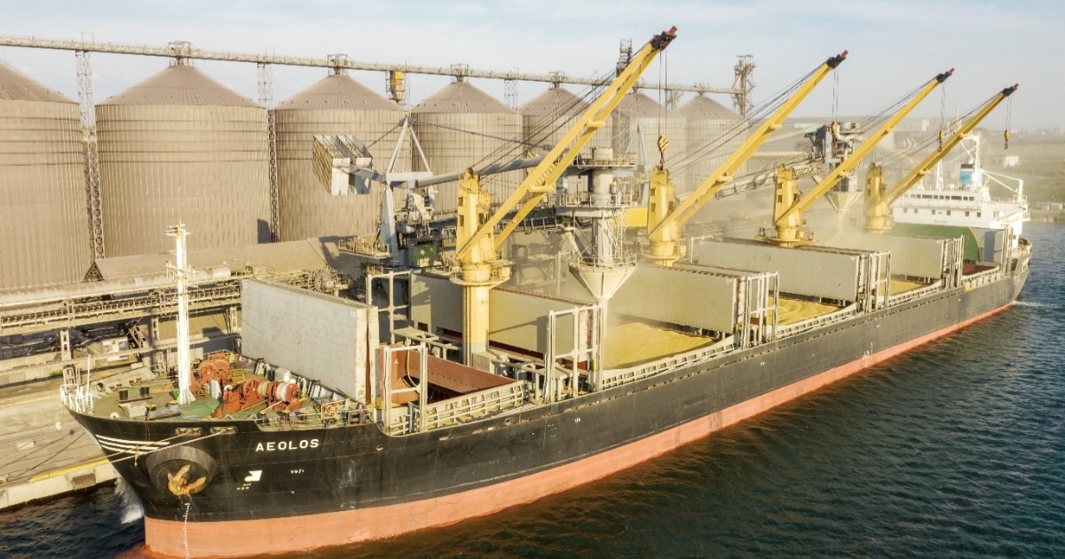 Imaginea prezintă o navă cargo de mari dimensiuni ancorată într-un port industrial lângă un rând de silozuri, în timp ce este încărcată cu cereale.