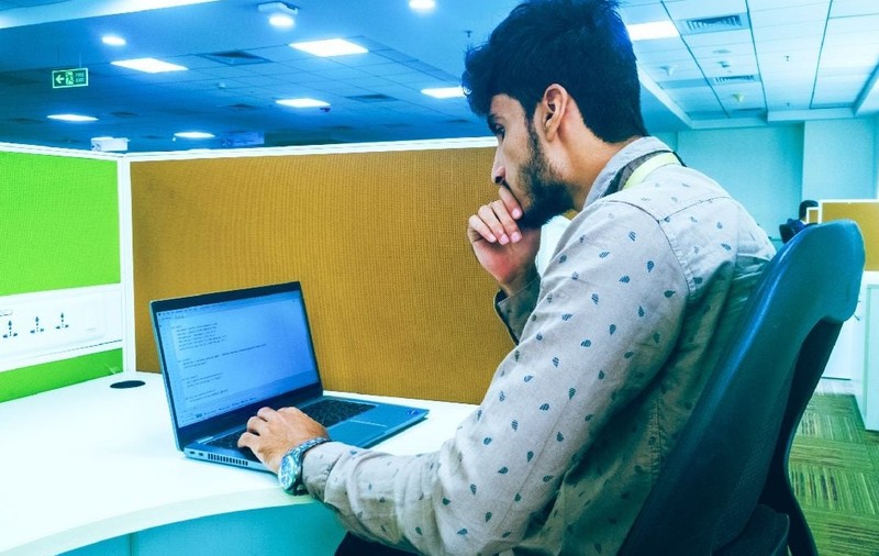 Imaginea surprinde un tânăr concentrat la locul de muncă dintr-un birou, stând la un computer.