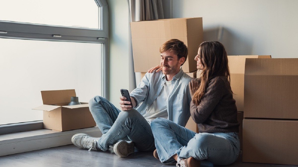 Ghid pentru mutarea în casă nouă: Checklist complet - Imagine cu un bărbat şi o femeie, uitâtndu-se pe un smartphone, într-o cameră fără mobilă şi având în spate cutii mari de carton