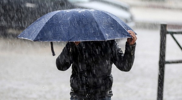 O persoană îmbrăcată într-o geacă neagră care se adăpostește sub o umbrelă albastră, ținând-o strâns cu ambele mâini în timpul unei ploi torențiale.