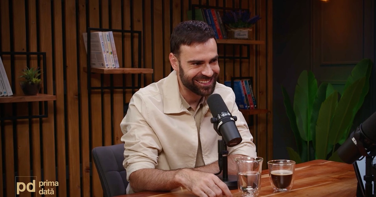 Denis Hanganu, în timpul unui podcast. E îmbrăcat cu o cămașă albă, stă la o masă și are în fața lui un microfon.