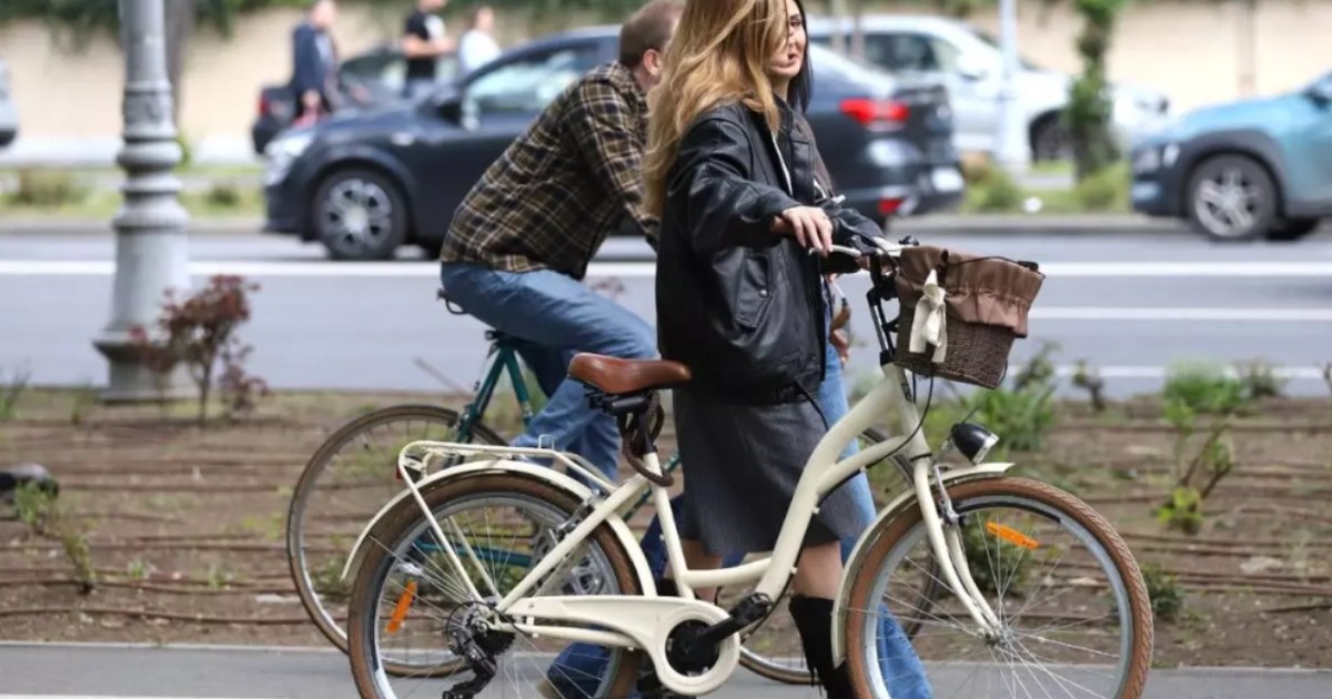 În imagine sunt două persoane care se plimbă cu bicicleta pe o alee din oraș, una dintre ele mergând pe lângă bicicletă, iar cealaltă pedalând.