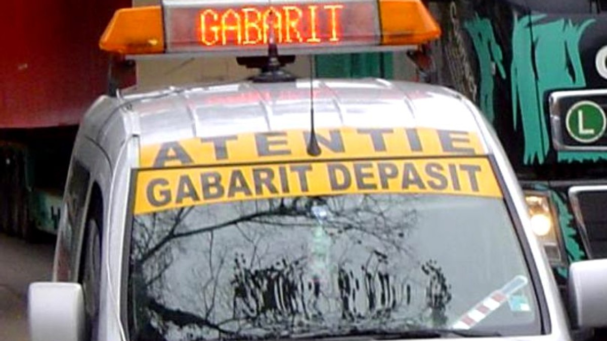 Patru transporturi agabaritice vor traversa Romania. Mașină care înoțește un convoi de transport agabaritic, pe care scrie "Atenție, gabarit depășit!"