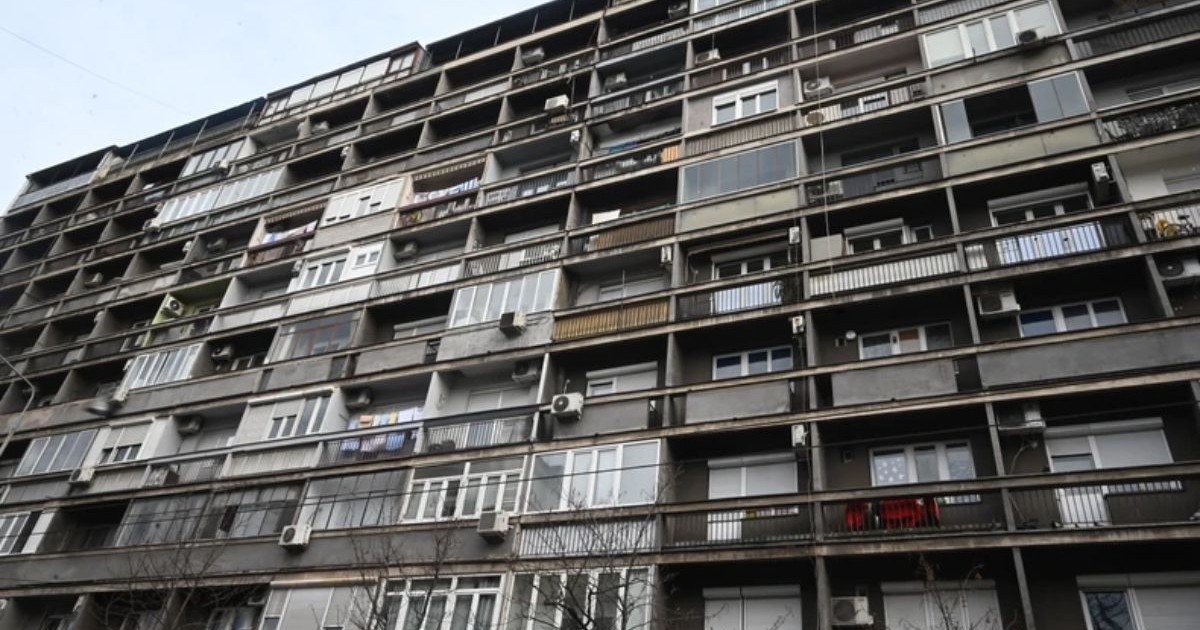 Un bloc vechi, fotografiat din față, cu balcoane de diferite culori - alb, gri și negru, cu cerul albastru deasupra