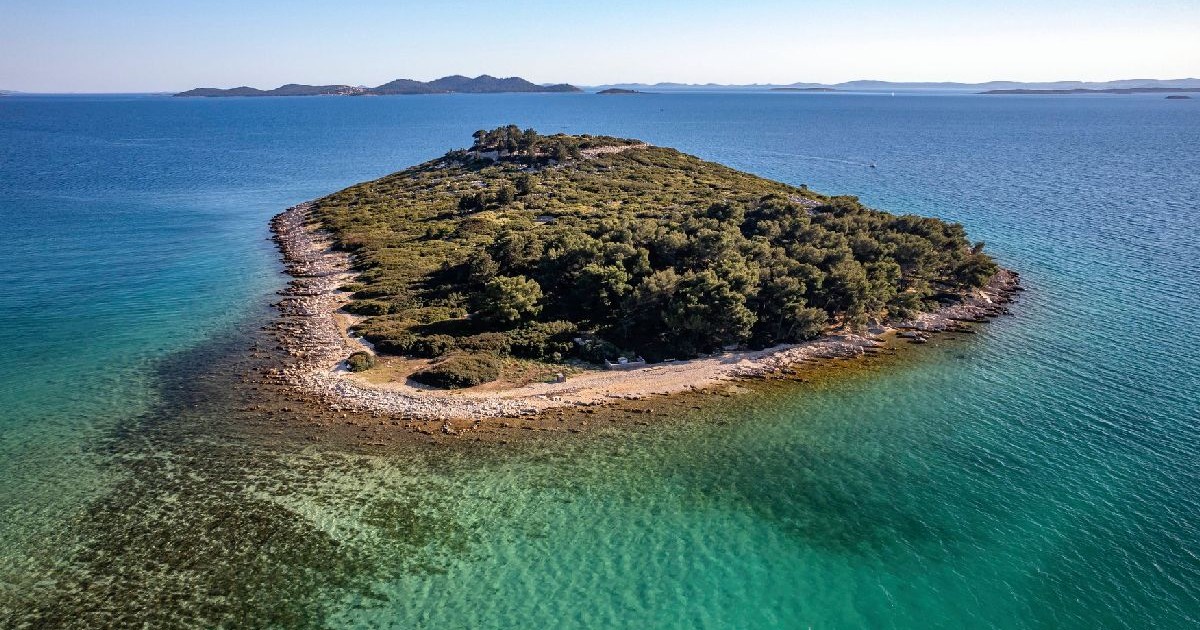 Fotografie aeriană a unei insule mici, luxuriante și verzi, situată într-o mare turcoaz. Insula, înconjurată de ape limpezi și puțin adânci, are o vegetație densă și un țărm stâncos. Cerul este senin și albastru.