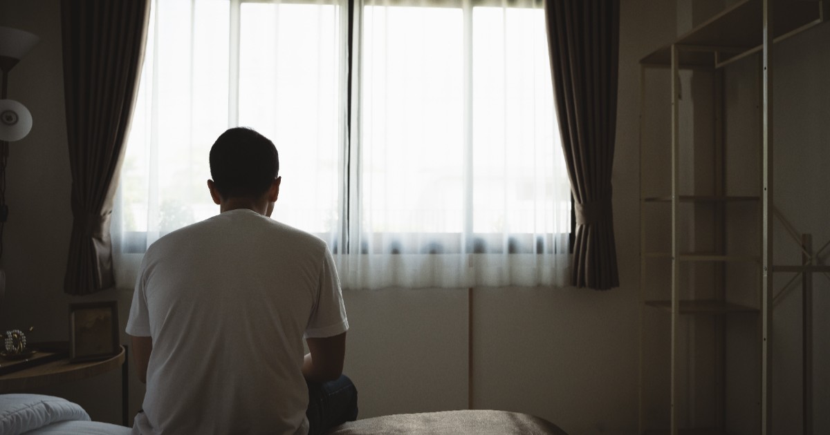 Imaginea arată un bărbat așezat pe marginea unui pat, cu spatele la cameră. Poartă un tricou alb și pare să privească spre fereastra din fața sa, prin care intră lumină difuză.