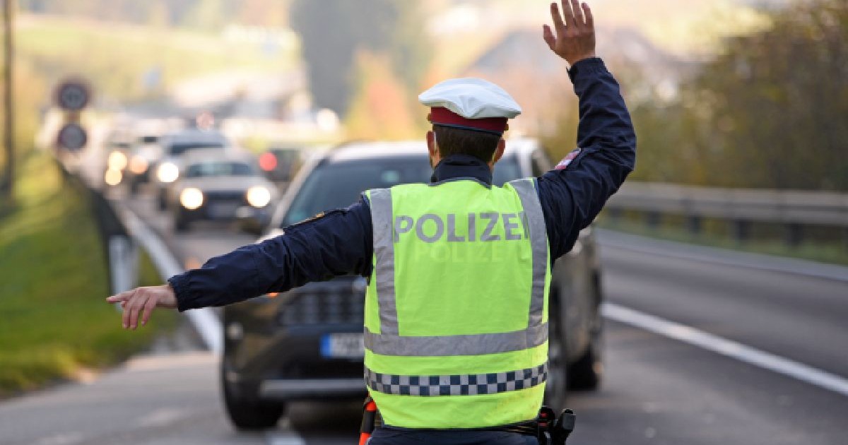 Imaginea surprinde un ofițer de poliție din spate, purtând o vestă reflectorizantă galbenă cu inscripția „POLIZEI”, în timp ce dirijează traficul pe o șosea.