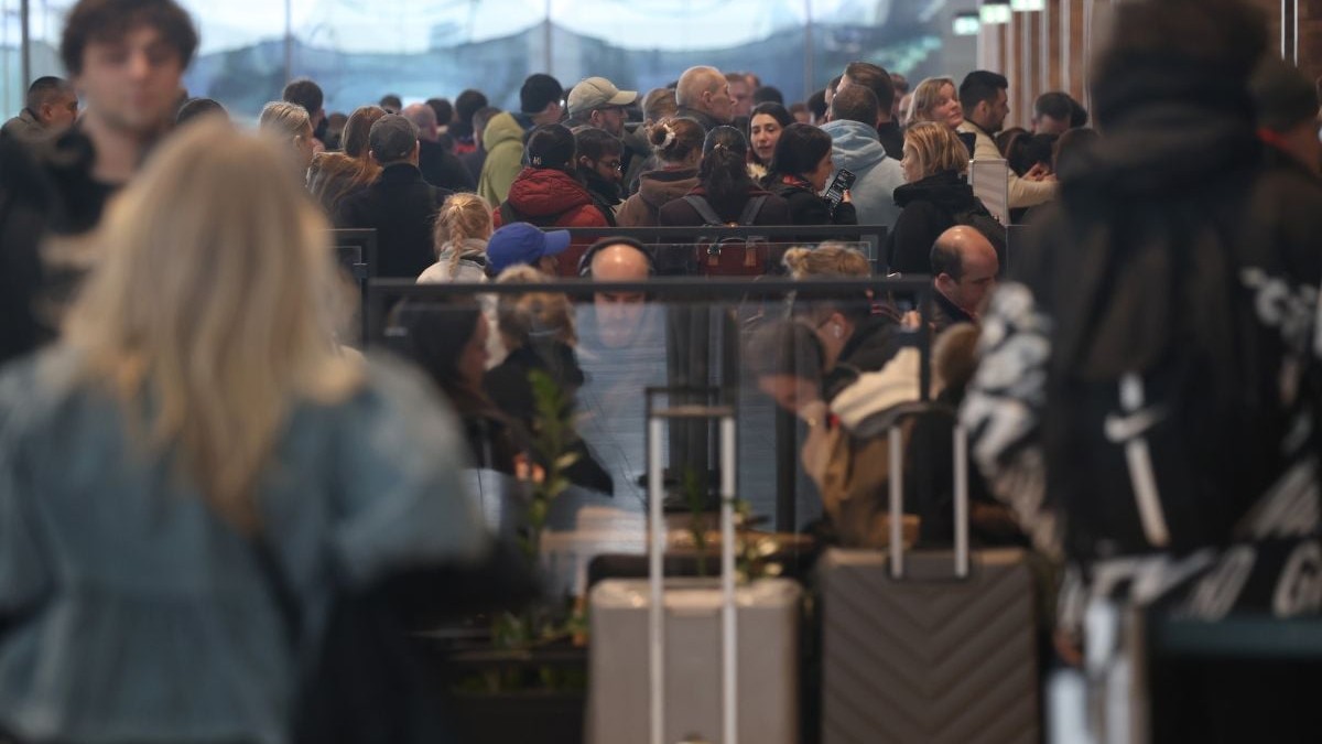 interiorul unui terminal extrem de aglomerat, unde zeci de călători stau foarte apropiați unii de alții, formând o masă densă de oameni. În prim-plan, ușor scoase din claritate, apar siluetele unor persoane și două valize cu mânere telescopice ridicate, indicând așteptarea lentă la o coadă. Persoanele din planul mijlociu și îndepărtat poartă haine specifice sezonului rece sau călătoriilor, cum ar fi geci voluminoase, șepci, glugi și căști audio, iar o parte dintre ei privesc concentrat în ecranele telefoanelor mobile. Printre mulțime se pot distinge câteva panouri despărțitoare din sticlă care fragmentează și organizează rândurile de oameni, subliniind senzația unui spațiu închis, plin până la refuz, surprins într-un moment de așteptare statică.
