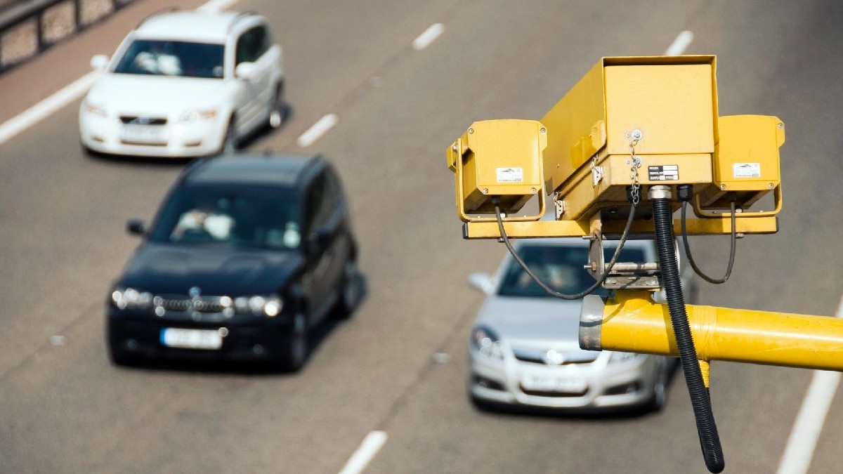 Fotografie cu o autostradă pe care se află trei mașini și o cameră de supraveghere a traficului galbenă montată pe un stâlp.