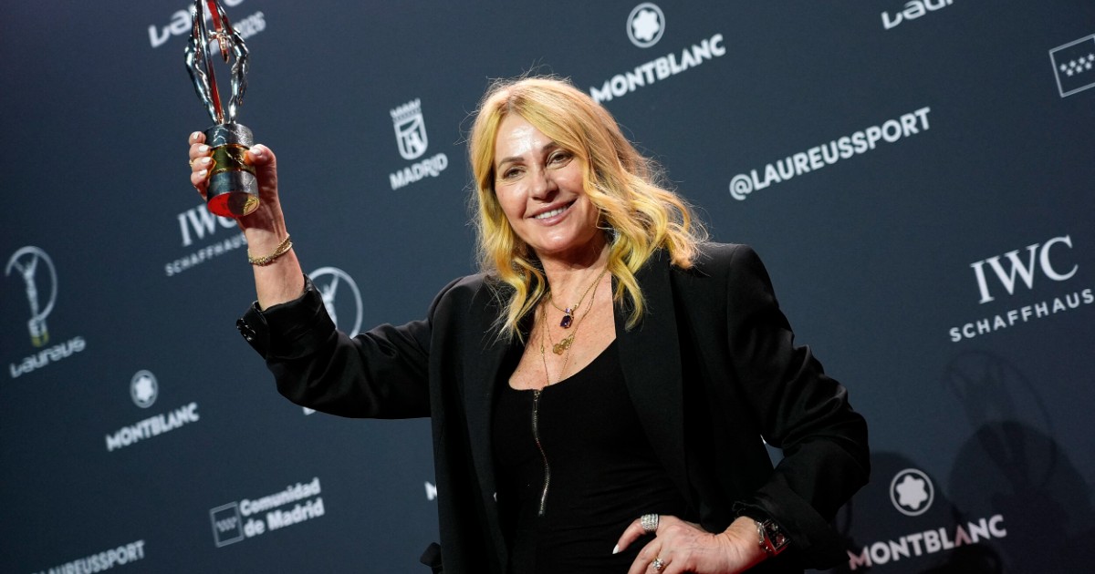 Nadia Comăneci apare zâmbitoare pe covorul roșu, ținând în mână trofeul primit la Gala Laureus, într-o ținută elegantă de culoare neagră. În fundal se observă panoul oficial al evenimentului, cu logo-urile sponsorilor și ale galei.