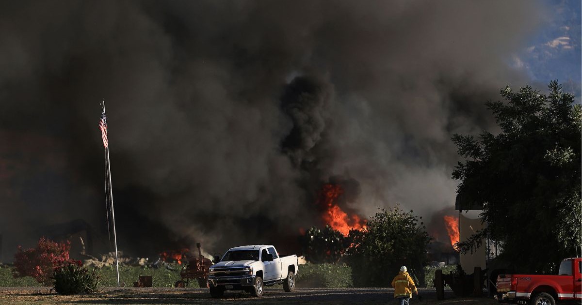 incendiu de vegetație care degază fum negru și gros. în partea de jos a imaginii se vede o pajiște verde, cu un pompier îmbrăcat în echipament galben, și două mașini pick-up mari. în spatele mașinilor se văd flăcări de culoare roșu aprins. în stânga imaginii se observă un catarg pe care este prins steagul SUA, care stă culcat în jos
