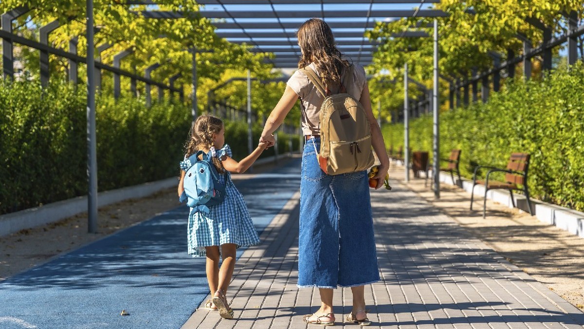 Mama și fiica ținându-se de mână și mergând împreună pe o alee pavată, cu rucsacurile în spate, într-o zi însorită.