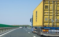 Imaginea surprinde un camion albastru ce transportă un container galben, rulând pe o autostradă alături de alte autoturisme, sub un cer senin.