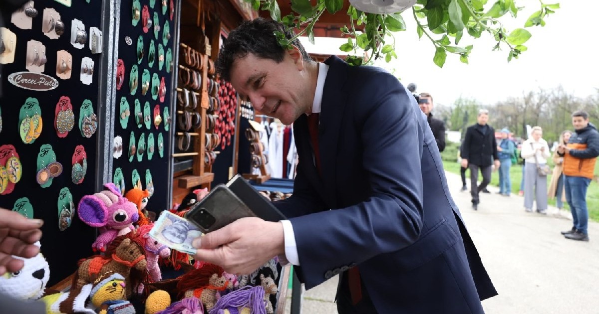 Nicușor Dan, îmbrăcat într-un costum negru, cu bani în mână, vrea să cumpere ceva de la o tarabă