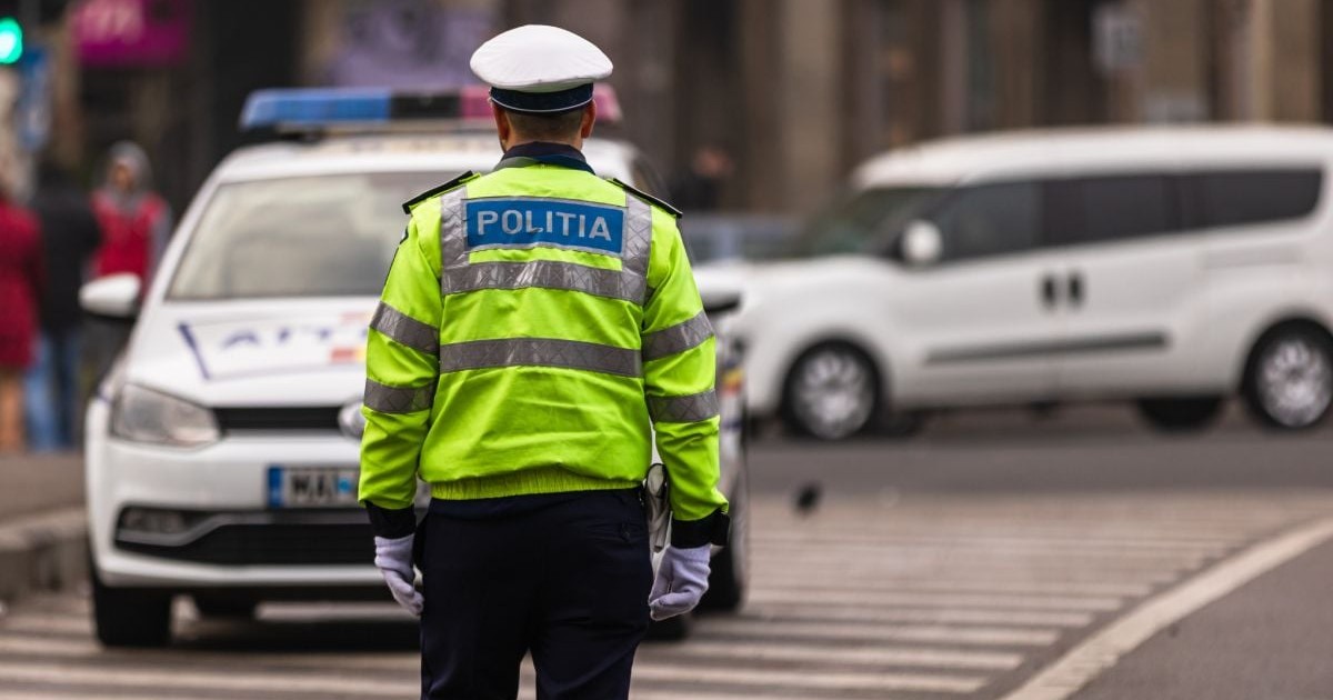 Un polițist de la Rutieră, îmbrăcat cu o geacă de culoare verde, pe stradă, iar pe fundal o mașină de Poliție în blur