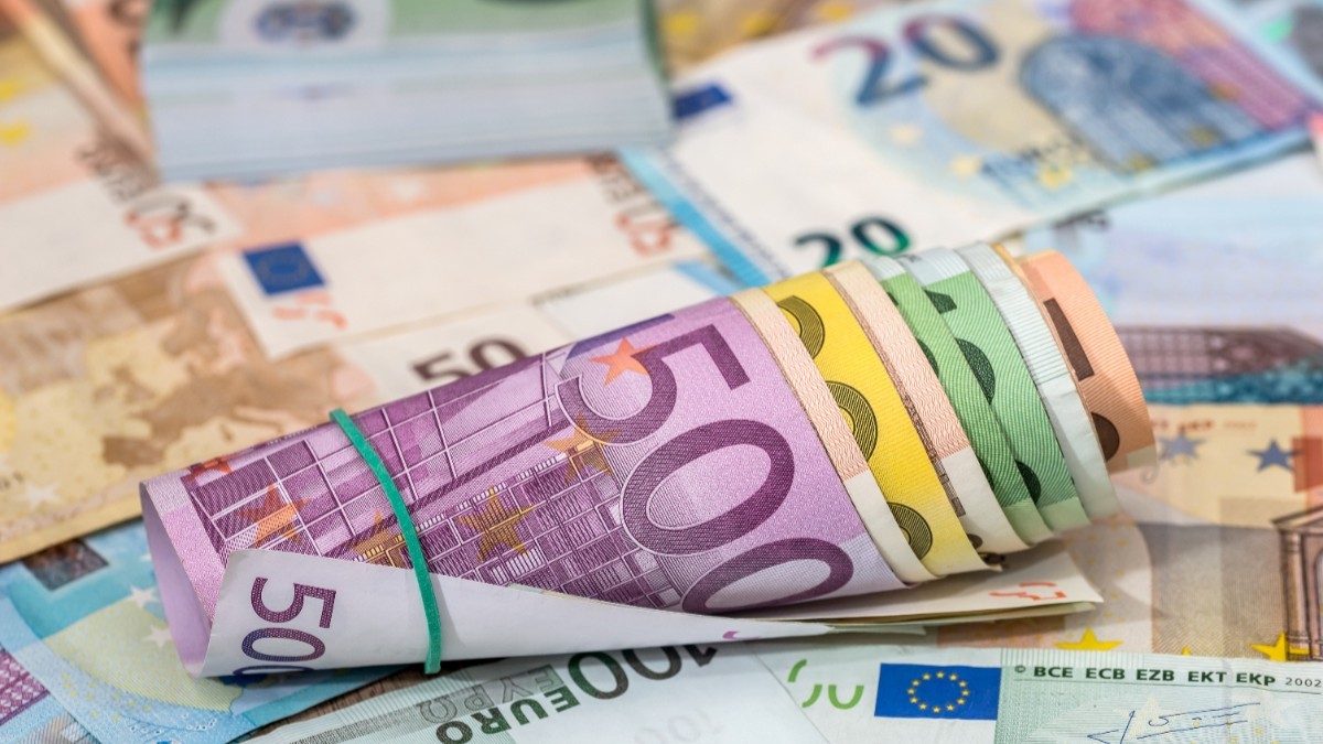 Sul de bancnote euro cu valori diferite legate cu un elastic, așezat pe un fundal acoperit de numeroase alte bancnote euro împrăștiate