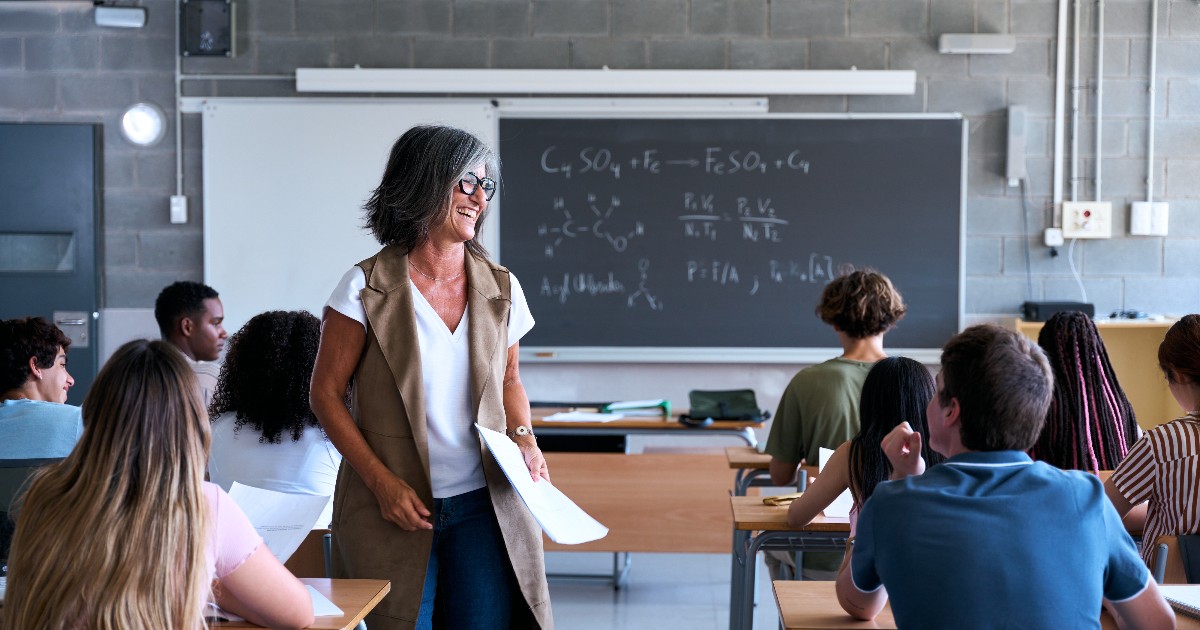 O profesoară zâmbitoare interacționează cu elevii într-o sală de clasă luminoasă, creând o atmosferă relaxată și deschisă. Imaginea surprinde un moment de implicare activă, în care elevii par atenți și interesați de desfășurarea orei.