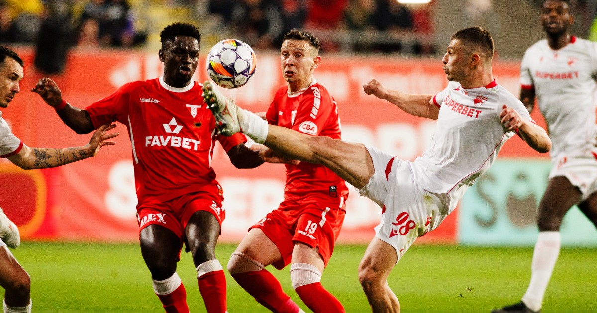 Imaginea surprinde un moment intens de luptă pentru minge în timpul unui meci de fotbal, cu mai mulți jucători de la Dinamo (în echipament roșu) și echipa adversă (în echipament alb) grupați în jurul mingii în aer. Unul dintre jucătorii în alb, cu numărul 98, execută o lovitură de picior înaltă, având piciorul întins la nivelul capului adversarului, pe un fundal marcat de o bandă de publicitate roșie și gazon verde.