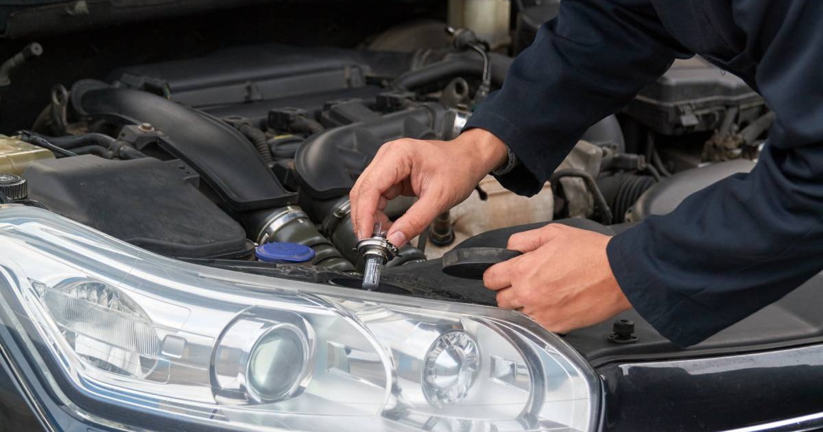 mecanic auto in dreptul unei masini care are capota deschisa. in mana tine un bec nou in dreptul farului