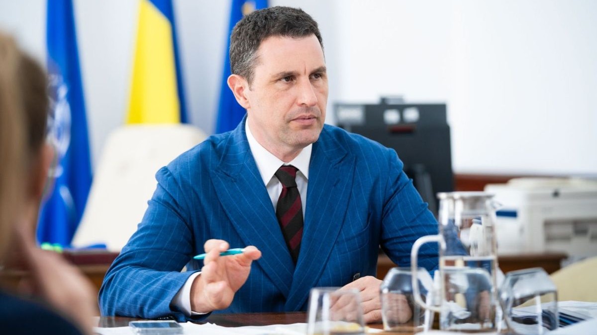 Ministrul interimar al Finanțelor, Tanczos Barna, îmbrăcat la costum, stă la o masă unde vorbește și explică dând din mâna stângă, în care are și un pix, iar în fața lui sunt pahare cu apă și în spate se văd steagul României, UE și al NATO