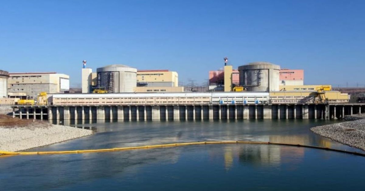Imagine cu Nuclearelectrica, se vad mai multe cladiri in spate, cu un baraj construit in față și apă, apa este traversată de un furtun