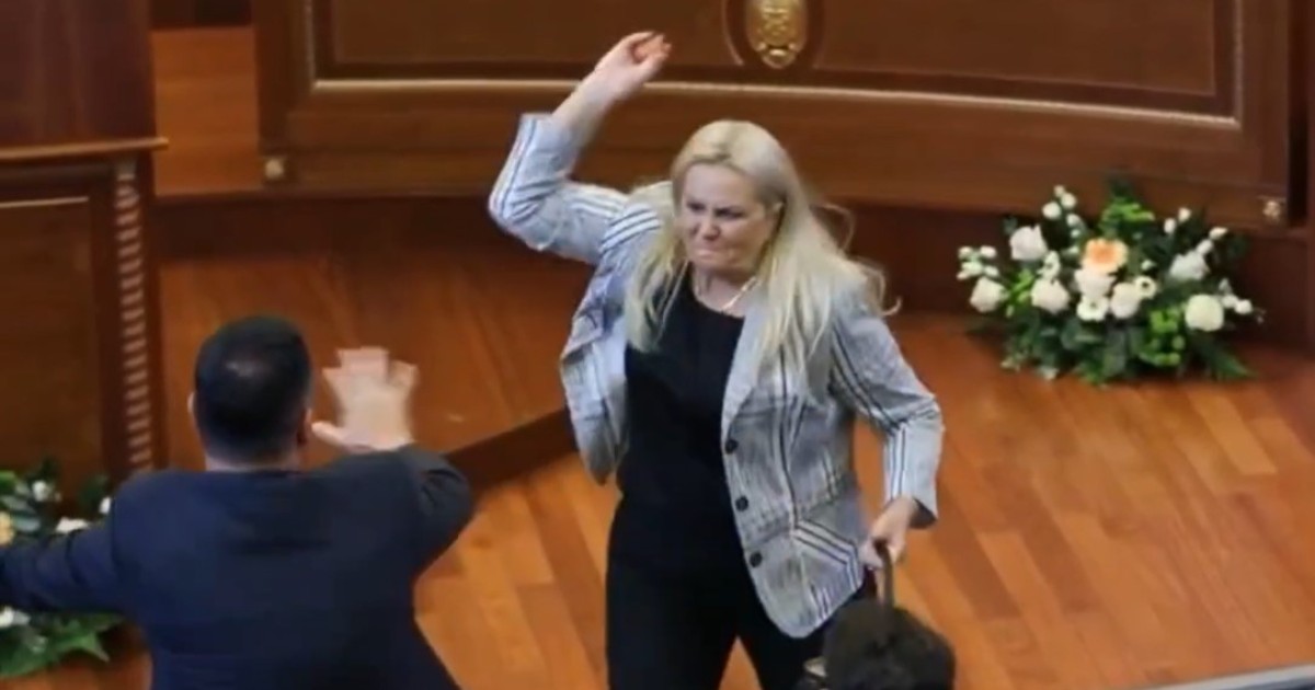 Imaginea prezintă o femeie blondă într-un sacou care ridică mâna pentru a arunca un obiect într-un bărbat dintr-o sală de parlament, în timp ce acesta încearcă să se apere.