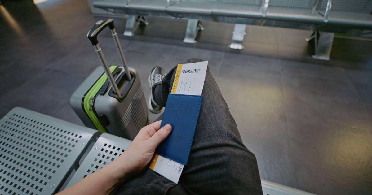 Calator sta asezat pe o banca in aeroport, picior peste picior, cu pasaportul si biletul de avion in mana stanga și un troller langa el