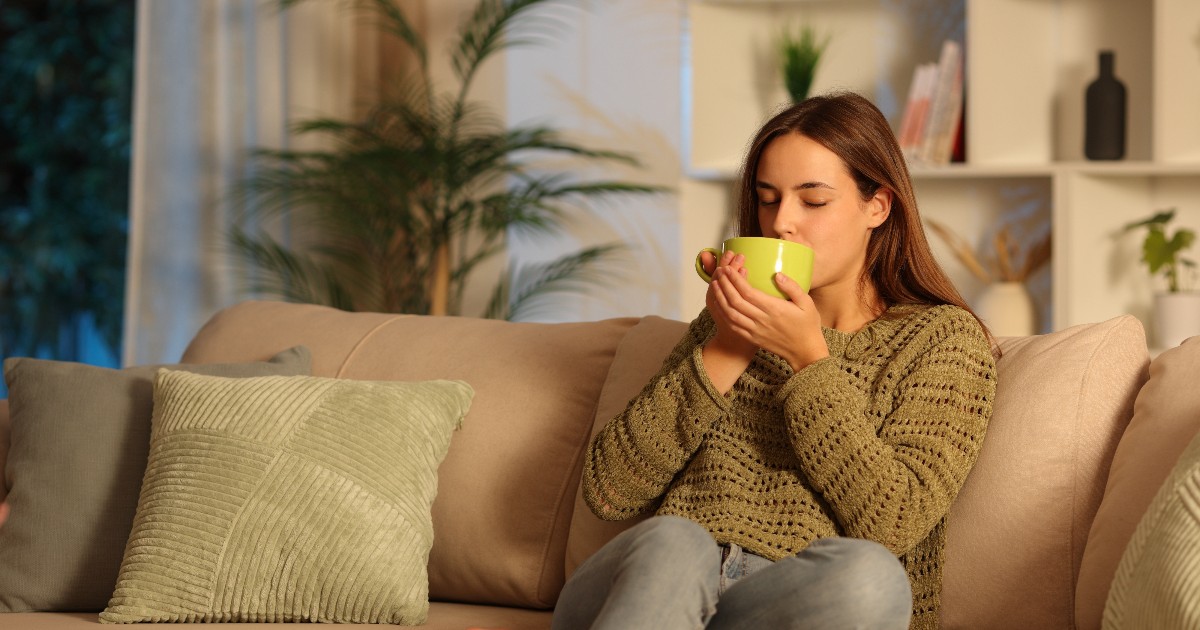 O femeie stă relaxată pe canapea, într-un living confortabil, și bea o băutură caldă dintr-o cană. Atmosfera este liniștită, sugerând un moment de relaxare de seară.