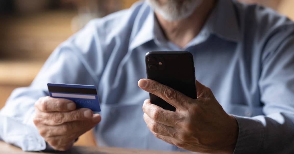 Cadru cu un barbat in varsta, in camasa albastra, asezat la o masa, intr-o mana tine un telefon, in cealalta un card de dredit. Conceptul de fraudă telefonică