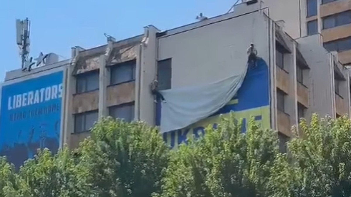 În imagine se vede o clădire din centrul orașului, pe fațada căreia este afișat un steag ucrainean de mari dimensiuni. Steagul este surprins în timp ce este coborât de pe clădire, în timp ce în partea laterală se poate observa un panou albastru cu mesajul „Liberators”.