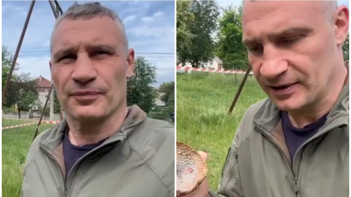 În imagine este Vitali Klitschko, fotografiat din față, purtând o geacă de culoare kaki și un tricou închis la culoare. Fundalul arată prezența unor case, copaci și o zonă împrejmuită cu bandă de avertizare.