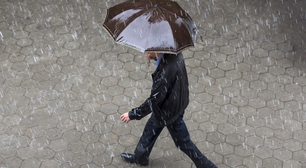 Un bărbat îmbrăcat într-o geacă neagră și pantaloni închiși la culoare, mergând pe un trotuar pavat în timp ce ține o umbrelă maro deasupra capului, pe o ploaie torențială.